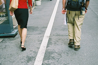 A white line divides the sidewalk. A woman walks on the left and a man on the right.